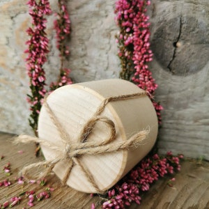 May include: A round wooden box with a natural finish, tied with twine. The box is surrounded by pink heather flowers on a wooden surface.
