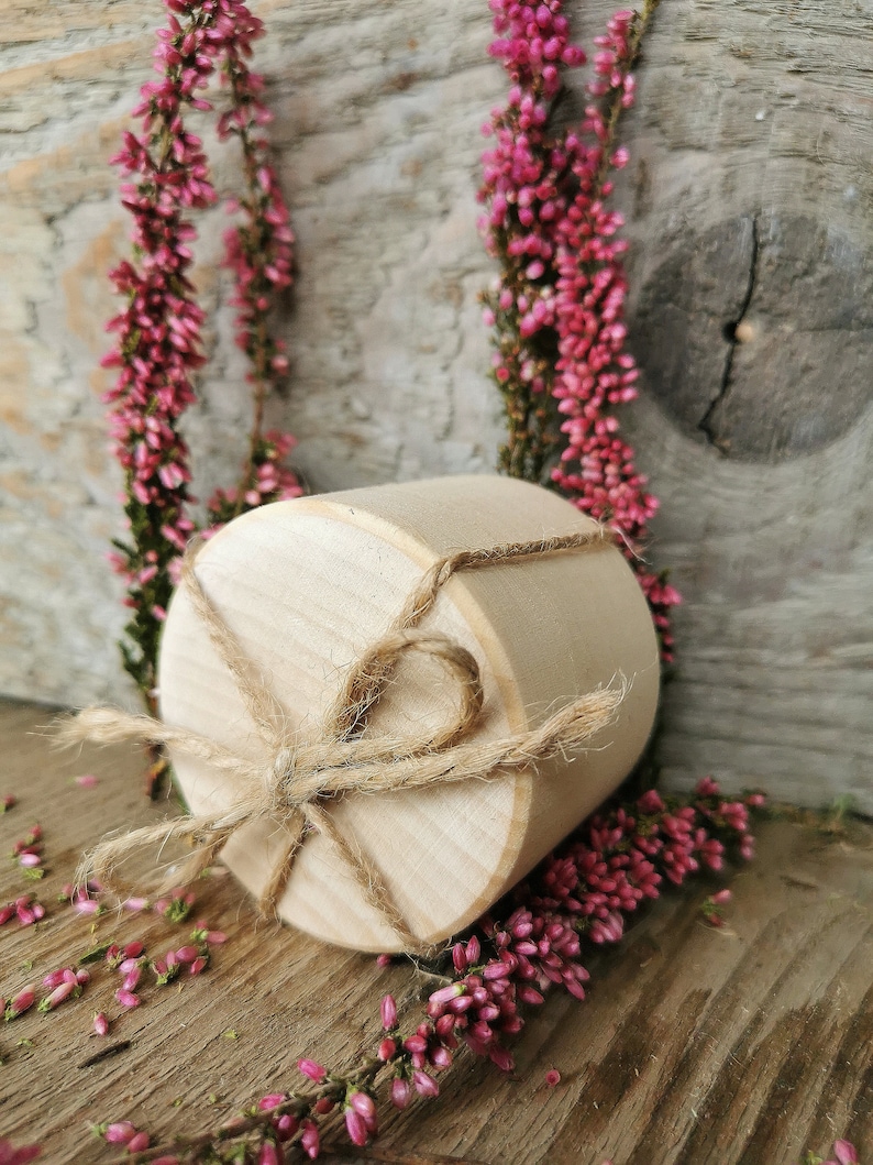 May include: A round wooden box with a natural finish, tied with twine. The box is surrounded by pink heather flowers on a wooden surface.