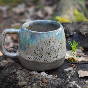 May include: A ceramic mug with a rounded shape and a thick handle. The upper portion features a glossy blue glaze that drips over a speckled off-white middle section, while the base is a solid, unglazed matte grey clay texture.