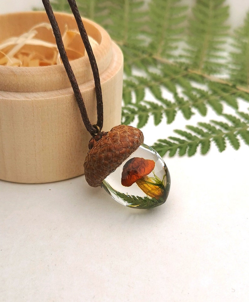 May include: A brown acorn pendant necklace with a clear resin dome containing a small orange mushroom and green foliage.
