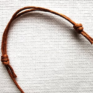 May include: Close-up of a brown leather cord with a knot tied in it. The cord is on a white fabric background.