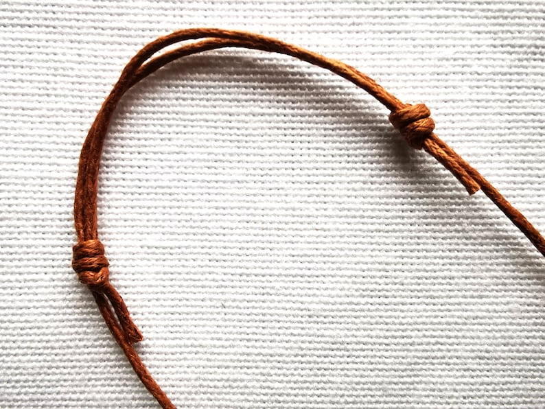 May include: Close-up of a brown leather cord with a knot tied in it. The cord is on a white fabric background.