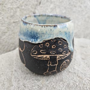 Può includere: Una tazza in ceramica fatta a mano con un esterno nero con un design a fungo intagliato. Il bordo è smaltato con un colore blu e bianco, creando un contrasto unico. La tazza è progettata per le bevande.