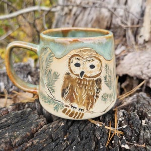 Puede incluir: Una taza de cerámica con base beige claro y borde esmaltado en verde. La taza presenta un búho marrón tallado y pintado a mano posado sobre una rama de pino. El asa es curva y combina con el esmalte verde del borde superior.