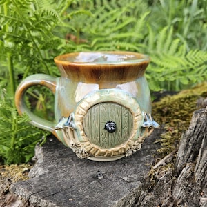 May include: A ceramic mug with a glossy glaze in shades of brown and sage green. The exterior features a sculpted circular door with a dark knob, flanked by small mushroom-like details and textured foliage elements at the base of the door.