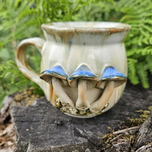 Könnte beinhalten: Ein Keramikbecher mit einer glänzenden cremefarbenen und braunen Glasur, der mit drei skulpturalen Pilzen mit leuchtend blauen Kappen an der Außenseite verziert ist. Der Becher hat einen bauchigen Körper, einen geschwungenen Henkel und einen strukturierten Fuß.