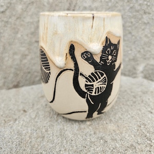 May include: A cream-colored ceramic tumbler with a black cat design. The cat is depicted playing with a ball of yarn. The tumbler has a unique, textured appearance, with a light brown, dripped effect near the rim.