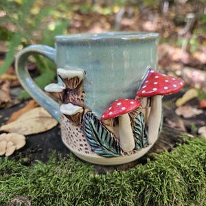 Puede incluir: Una taza de cerámica con esmalte azul claro que presenta detalles esculpidos en tres dimensiones. El exterior muestra setas rojas con puntos blancos, pequeños hongos blancos y hojas verdes sobre una superficie texturizada.