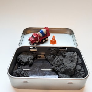 May include: A red toy cement mixer truck and an orange traffic cone are in a silver tin with black sand. The truck is driving on a black and white road.