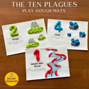 May include: Play dough mats with the text "The Ten Plagues". The mats feature colorful play dough creations of frogs, flies, and a river of blood. The number 2, 4, and 1 are also displayed. The mats are on a wooden surface.