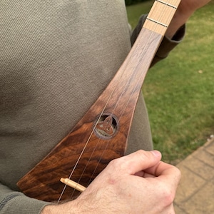 May include: A wooden, triangular-shaped musical instrument with strings. The instrument has a dark brown body with a lighter wood neck and fretboard. A Celtic knot design is carved into the body. The instrument is held by a person.