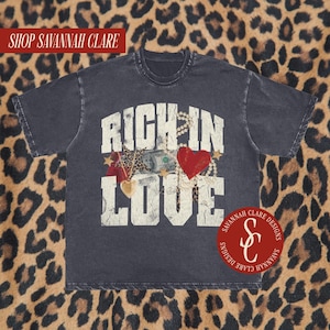 May include: A black t-shirt with a distressed vintage look featuring the text "RICH IN LOVE" in white with a graphic of a heart, money, and jewellery. The shirt also has a small logo with the text "SAVANNAH CLARE DESIGNS" and the initials "S&C" in red.