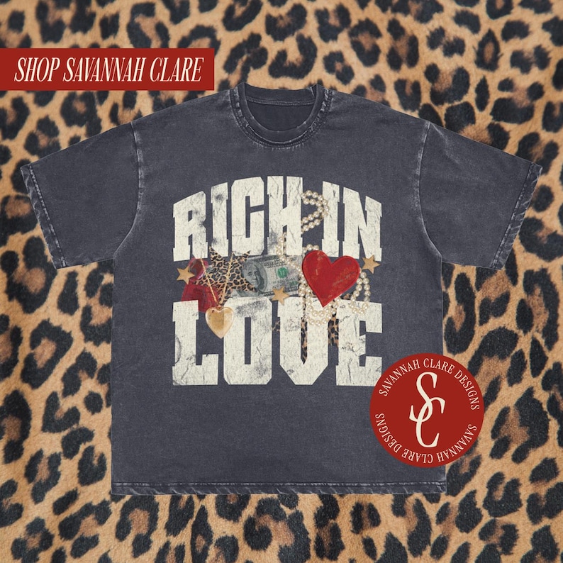 May include: A black t-shirt with a distressed vintage look featuring the text "RICH IN LOVE" in white with a graphic of a heart, money, and jewellery. The shirt also has a small logo with the text "SAVANNAH CLARE DESIGNS" and the initials "S&C" in red.