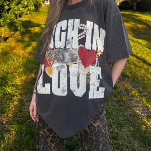 May include: An oversized dark grey t-shirt with the text "Rich in Love" printed on the front. The text is surrounded by a pearl necklace, a gold chain, a red heart, and a dollar bill.