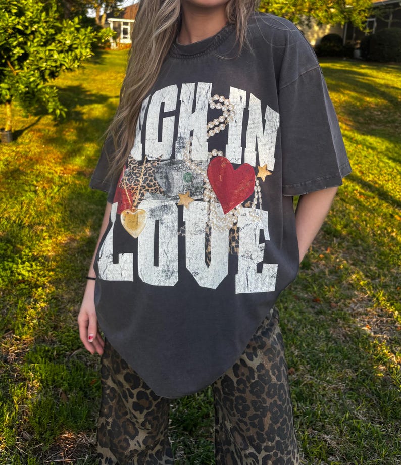 May include: An oversized dark grey t-shirt with the text "Rich in Love" printed on the front. The text is surrounded by a pearl necklace, a gold chain, a red heart, and a dollar bill.