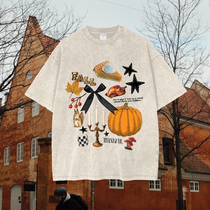 May include: Cream-coloured t-shirt with a fall-themed graphic. The design includes a pumpkin, pie, stars, a squirrel, and the word "FALL". A black bow and the word "THANKFUL" are also featured.