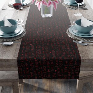 May include: A black table runner with a repeating red script pattern of words like "love", "adore", and "forever". The runner is placed on a wooden table set for a meal, with plates, glasses, and flowers.