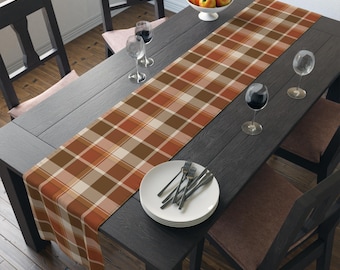 May include: A plaid table runner in shades of brown, orange, and cream, placed on a dark wooden table. The table is set with plates, silverware, wine glasses, and a bowl of fruit. The runner adds a touch of warmth and style to the dining setting.