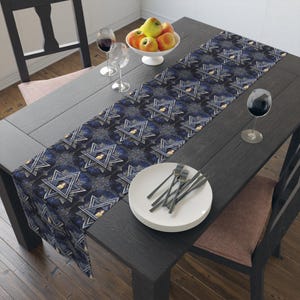 May include: A dark blue table runner with a repeating pattern of Star of David symbols. The runner is placed on a dark wooden table, with a white bowl of fruit, wine glasses, and a plate with forks.