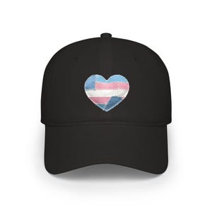 Op de afbeelding: Zwarte baseballcap met een hartvormige patch in de kleuren van de transgender pride vlag: lichtblauw, roze en wit. Het hart is gemaakt van gestikte stukken stof. De cap heeft een gebogen klep en een gestructureerde kroon.
