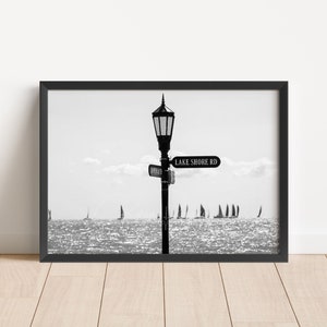 May include: Black and white photograph of a street sign that reads "Lake Shore Rd" with a lamppost in the center of the image. The sign is pointing to the right. There are sailboats in the distance.