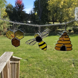 May include: A stained glass bee, honeycomb, and beehive hanging from a silver chain. The bee is yellow and black with clear wings. The honeycomb is yellow and brown. The beehive is yellow with a black entrance.