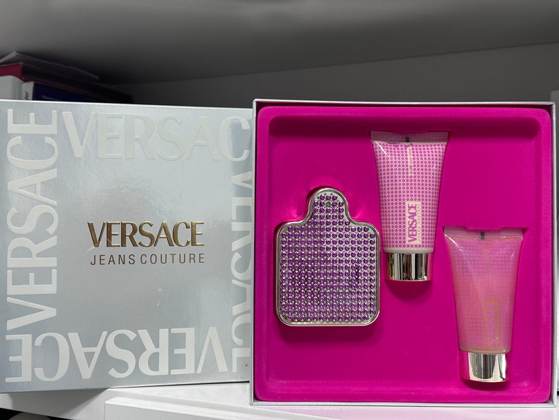 Op de afbeelding: Een roze cadeauset met een zilveren en paarse fles Versace Jeans Couture parfum, een roze lotionfles en een roze douchegel fles.