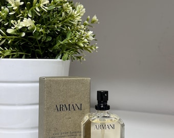 May include: A 1 fl oz bottle of Armani Eau Pour Homme cologne. The bottle is clear glass with a gold cap and a black spray nozzle. The bottle is sitting on a white surface with a green plant in the background.