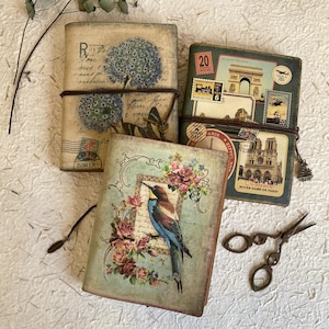 Op de afbeelding: Drie vintage dagboeken met verschillende ontwerpen. Het eerste dagboek heeft een bloemmotief met een blauwe bloem en bruine tekst. Het tweede dagboek heeft een bloemmotief met een vogel en roze bloemen. Het derde dagboek heeft een reisthema met postzegels en afbeeldingen van Parijs.