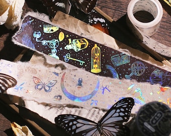 May include: Two rolls of iridescent holographic washi tape with different designs. One roll features a dark background with gold and silver accents, and the other roll features a light background with gold and silver accents. The designs include butterflies, moons, stars, and other celestial elements.