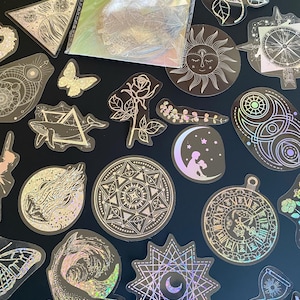 May include: A set of 21 holographic stickers featuring various celestial and nature-inspired designs. The stickers are iridescent and have a black background. Some of the designs include a sun, moon, stars, a whale, a butterfly, a rose, and a compass.