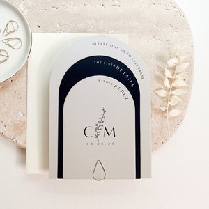 Puede incluir: Una tarjeta de invitación con un diseño arqueado, con el texto "PLEASE JOIN US TO CELEBRATE THE FINER DETAILS KINDLY REPLY". La tarjeta incluye iniciales y una fecha. Clips dorados y un plato blanco en el fondo.