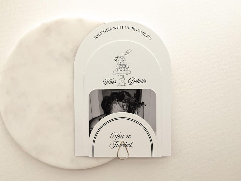 May include: White wedding invitation with a black and white photo of a couple kissing. The invitation has the text "Together with their families", "the Finer Details", and "You're Invited". A silver ring holds the invitation together.