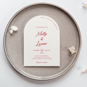 May include: A wedding invitation with a cream background and red text. The invitation is shaped like an arch and reads "Join us as we celebrate the wedding of Molly & Lenox, October 16, 2024, 4:00 PM, Riverside Meadows, 2436 Spring Trail Drive, Asheville, North Carolina 28801, Reception to follow."
