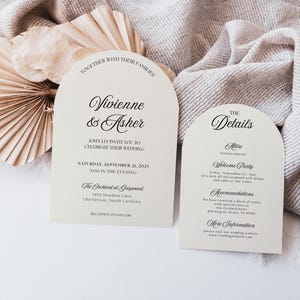 May include: Two ivory wedding invitation cards with arched tops. The left card reads "Vivienne & Asher" and the right card says "The Details". The cards are on a neutral-toned fabric with dried palm leaves.