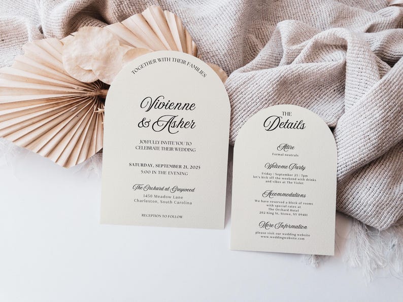 May include: Two ivory wedding invitation cards with arched tops. The left card reads "Vivienne & Asher" and the right card says "The Details". The cards are on a neutral-toned fabric with dried palm leaves.