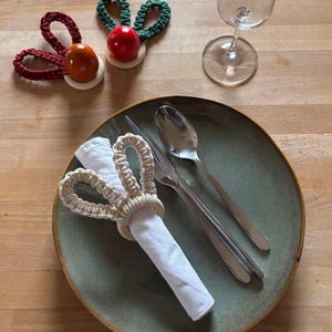 Op de afbeelding: Een groene, rode en bruine bunny servetring gemaakt van touw, een witte servet, een zilveren vork, een zilveren mes en een zilveren lepel op een groen bord.