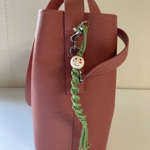 Puede incluir: Un bolso de color rosa polvoriento con un asa a juego y un llavero de macramé verde. El llavero presenta una cuenta de madera con una cara sonriente. El bolso tiene un diseño estructurado y una superficie texturizada.