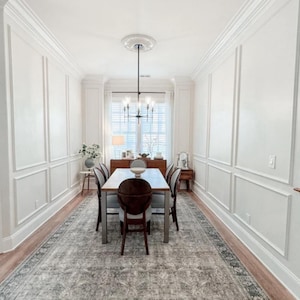 May include: A dining room featuring a rectangular wooden table with six chairs, a large area rug with a grey and white pattern, white walls with moulding, a black metal chandelier, and a wooden sideboard positioned against a window with blinds.