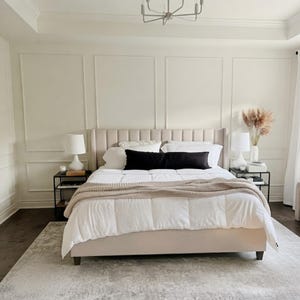 May include: A beige upholstered bed with a vertical channel tufted headboard, dressed in a white comforter, a long black lumbar pillow, and a chunky knit beige throw blanket. Two black metal nightstands hold white lamps and decorative items.