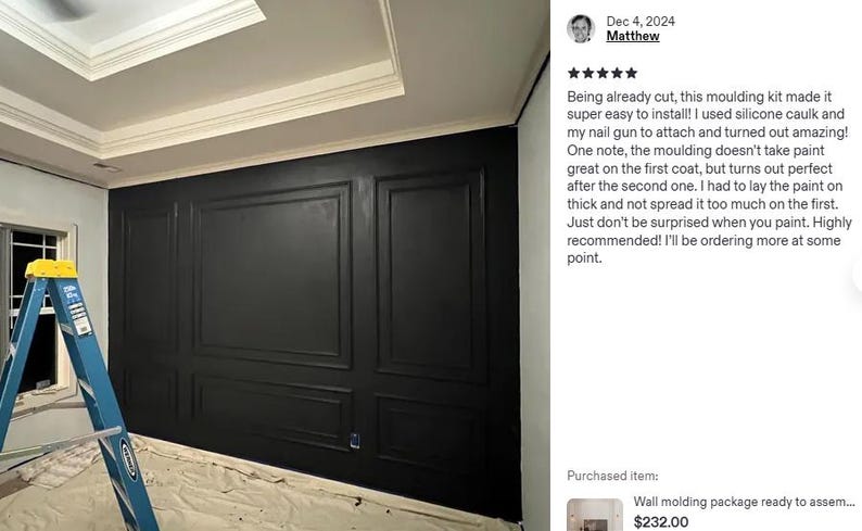 May include: A wall featuring black rectangular moulding panels installed in a grid pattern. A blue step ladder stands in the foreground. The wall is painted a solid matte black, creating a framed panel effect across the entire surface area.