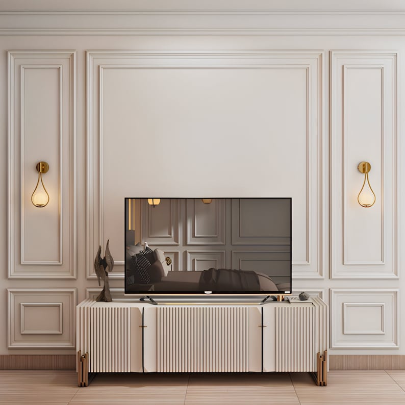 May include: A white entertainment center with a flat screen TV on top. The entertainment center has a gold metal frame and vertical slats. Two gold wall sconces with teardrop-shaped glass shades are on either side of the TV.