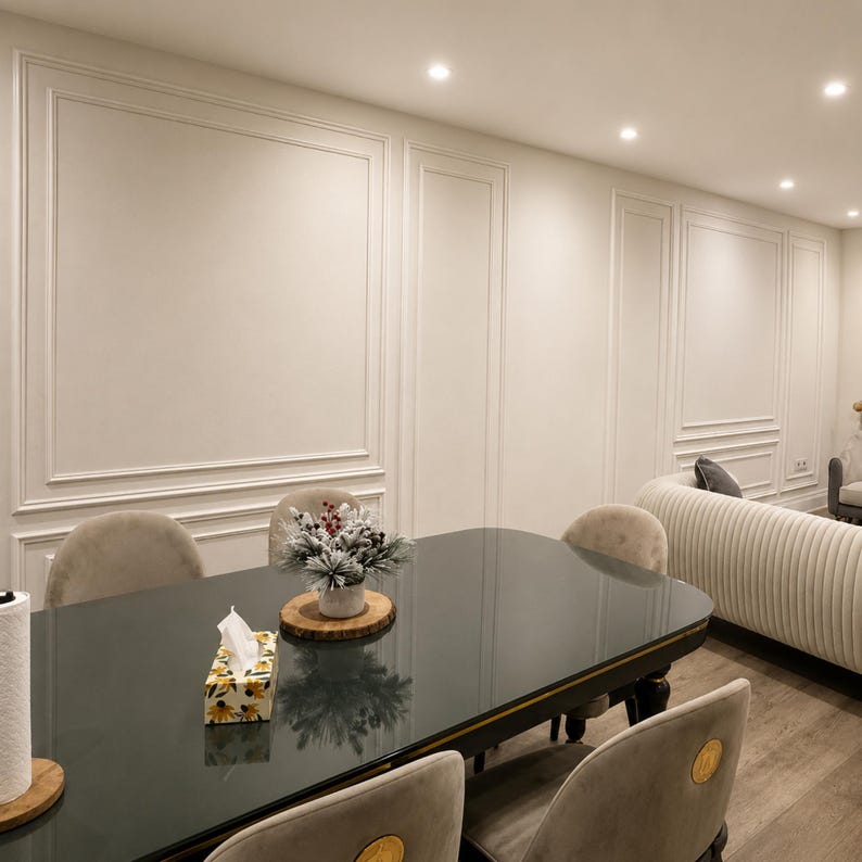 May include: A dark gray oval dining table with a gold trim edge, surrounded by beige velvet chairs. On the table sits a floral tissue box and a small decorative pine arrangement on a wooden base. The room features white walls with molding.