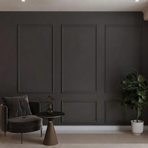 May include: A dark charcoal grey wall with rectangular moulding panels, a matching velvet armchair, a small round side table with a dark glass vase containing dried branches, and a green potted plant in a white ceramic pot.