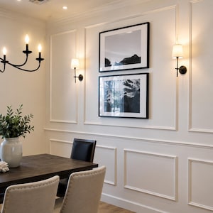May include: A dining room wall with white wainscoting features two stacked black-framed landscape photographs of mountains. Two wall-mounted sconces with white fabric shades flank the artwork, while a black chandelier hangs nearby.