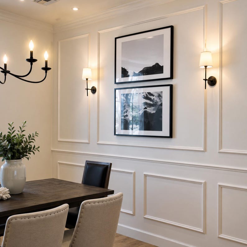 May include: A dining room wall with white wainscoting features two stacked black-framed landscape photographs of mountains. Two wall-mounted sconces with white fabric shades flank the artwork, while a black chandelier hangs nearby.