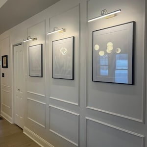 May include: A hallway with white paneling and three framed pictures. Each picture has a black frame and is illuminated by a gold-toned picture light. The wall paneling has a decorative design.