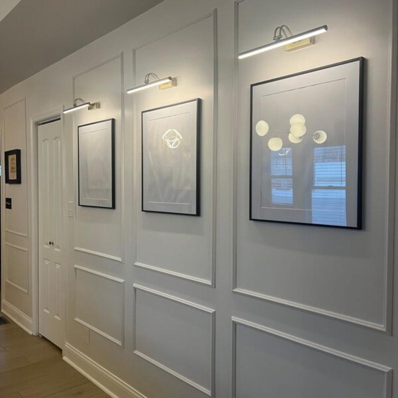 May include: A hallway with white paneling and three framed pictures. Each picture has a black frame and is illuminated by a gold-toned picture light. The wall paneling has a decorative design.