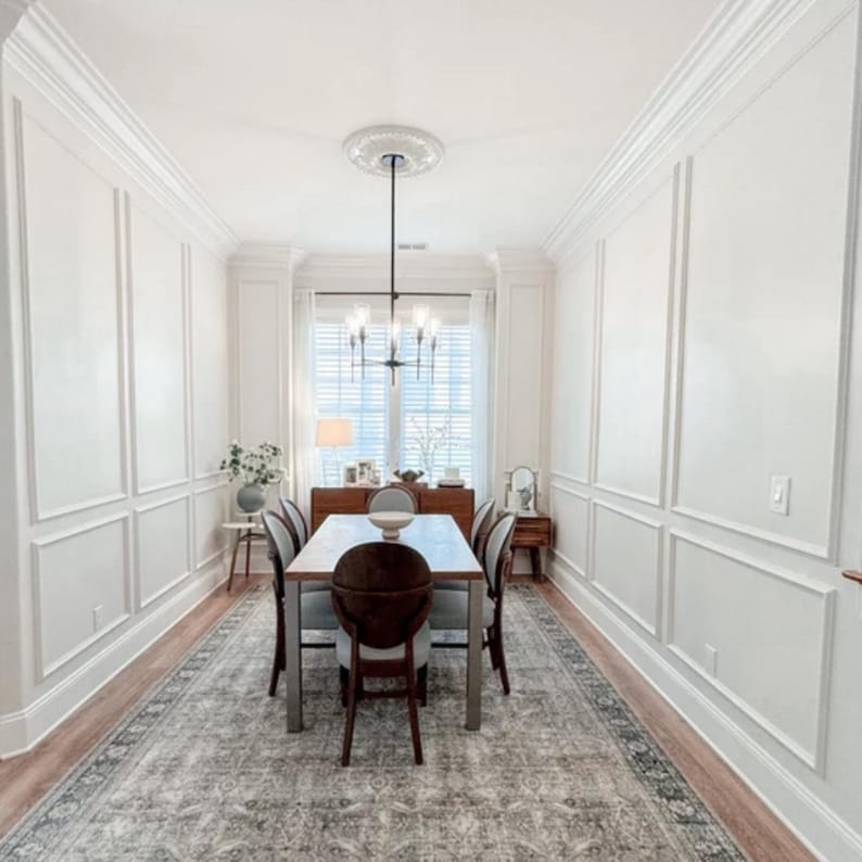 May include: A dining room featuring white walls with rectangular molding, a rectangular wooden table with six chairs, a dark metal chandelier with six lights, and a large gray patterned area rug covering the light wood floor.