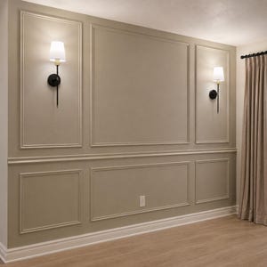 May include: A beige wall featuring decorative rectangular molding panels and two wall-mounted sconces with black metal stems and white fabric shades. The wall is accented by a horizontal chair rail, white baseboards, and light-colored wood flooring.